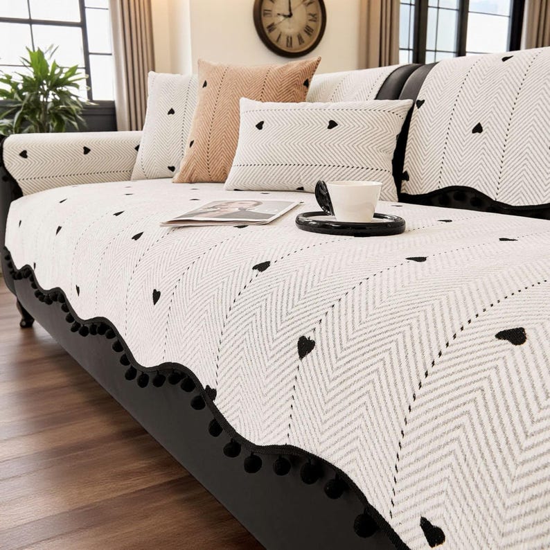 May include: A black sofa with a white and gray patterned cover featuring black heart accents. Decorative pillows in white, pink, and gray complement the sofa. A black trim with black pom-poms lines the bottom edge of the cover.