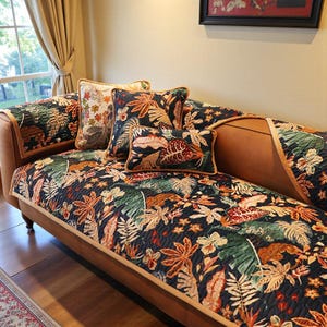 May include: A brown chaise lounge with a dark floral patterned cover and matching pillows. The pattern features green, orange, and cream-colored leaves and flowers on a dark background. The pillows have a gold trim.
