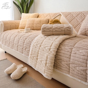 May include: A beige sofa with matching throw pillows and a blanket. The sofa and pillows have a cable knit design. A rolled-up blanket and a pair of fuzzy slippers are also visible. The text "HG Home Comfort" is in the upper left corner.