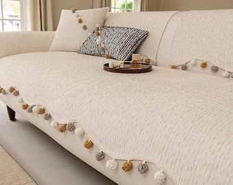 May include: Off-white sofa with a textured pattern, adorned with decorative pom-poms along the edges. Includes matching throw pillows, one with a black and white striped pattern. A small wooden tray with a glass bottle sits on the sofa.