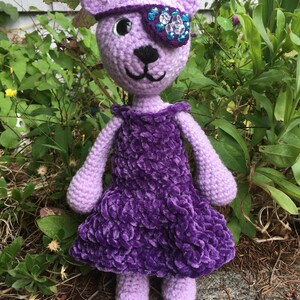May include: A purple crocheted teddy bear wearing a purple dress and a sparkly eye patch. The bear is standing on a bed of green leaves.