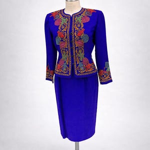 May include: A vibrant blue skirt suit with an embroidered jacket. The jacket features colorful floral and seashell designs in red, green, yellow, and purple. The skirt is a matching solid blue. The suit is displayed on a mannequin.