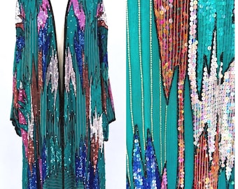 May include: A long, open-front jacket in teal green featuring intricate vertical patterns of multi-coloured sequins and beads. The garment displays jagged, flame-like shapes in shades of pink, blue, silver, and copper, with black trim along the edges.