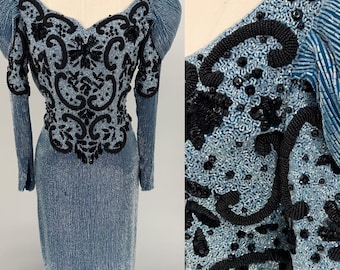 May include: A long, light blue formal dress with long sleeves and a V-neck. The bodice is embellished with intricate black beading in a floral pattern. The dress is displayed on a mannequin, showcasing its elegant design and vertical striped texture.