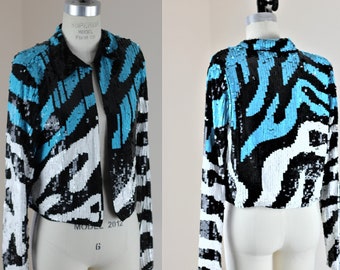 May include: A cropped sequined jacket with a black, white and turquoise abstract pattern. The jacket has a zip closure and long sleeves.