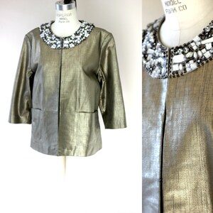 May include: A metallic gold jacket with a jewel-encrusted collar. The jacket has three-quarter sleeves and two front pockets. The collar is adorned with a mix of clear and black stones. The jacket is displayed on a mannequin.