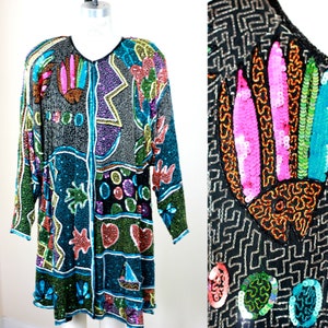 May include: A long-sleeved sequined tunic with a colorful abstract pattern. The tunic features a variety of shapes, including hearts, stars, and geometric designs. The sequins are in shades of blue, green, pink, purple, and orange.