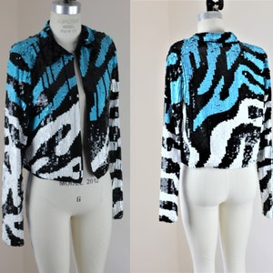 May include: A cropped sequined jacket with a black, white, and turquoise abstract pattern. The jacket has a zipper closure and long sleeves.