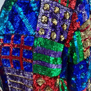 Puede incluir: Primer plano de una chaqueta cubierta de lentejuelas de colores. Las lentejuelas est&aacute;n dispuestas en un dise&ntilde;o de patchwork, con cuadrados y rect&aacute;ngulos azules, rojos, verdes, morados y dorados. Las lentejuelas son brillantes y reflejan la luz.