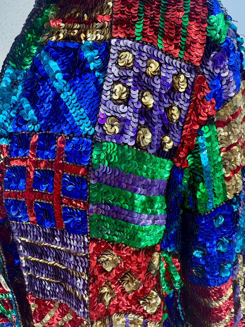 Puede incluir: Primer plano de una chaqueta cubierta de lentejuelas de colores. Las lentejuelas est&aacute;n dispuestas en un dise&ntilde;o de patchwork, con cuadrados y rect&aacute;ngulos azules, rojos, verdes, morados y dorados. Las lentejuelas son brillantes y reflejan la luz.