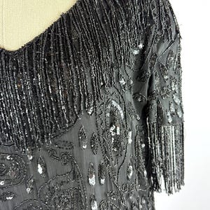 May include: A black, beaded dress with a scoop neckline and short sleeves. The dress features intricate sequin and bead embellishments, including fringe along the neckline and sleeves. The fabric has a paisley-like pattern.