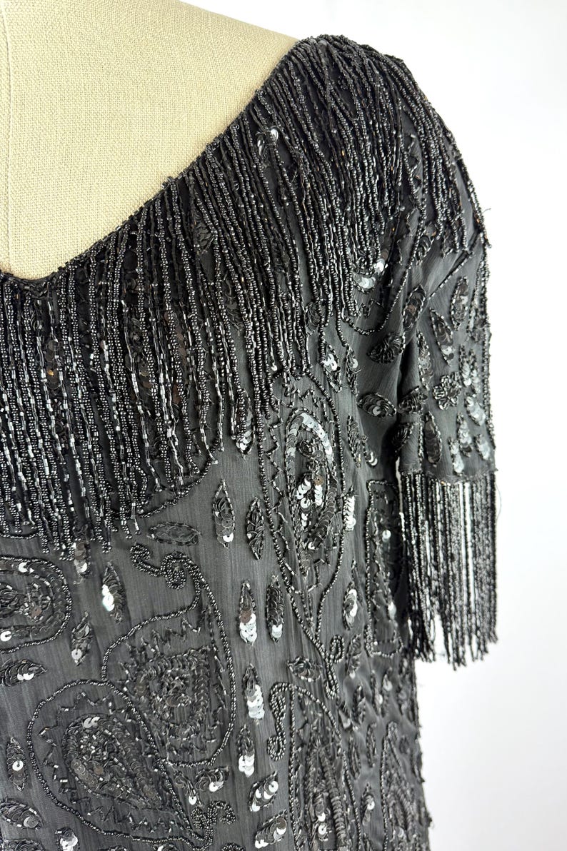 May include: A black, beaded dress with a scoop neckline and short sleeves. The dress features intricate sequin and bead embellishments, including fringe along the neckline and sleeves. The fabric has a paisley-like pattern.