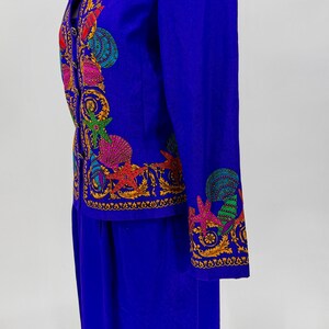 May include: A vibrant blue two-piece suit featuring intricate, colorful embroidered seashell and starfish designs. The jacket has long sleeves and a tailored fit, paired with a matching skirt. The embroidery includes shades of red, green, yellow, and pink.
