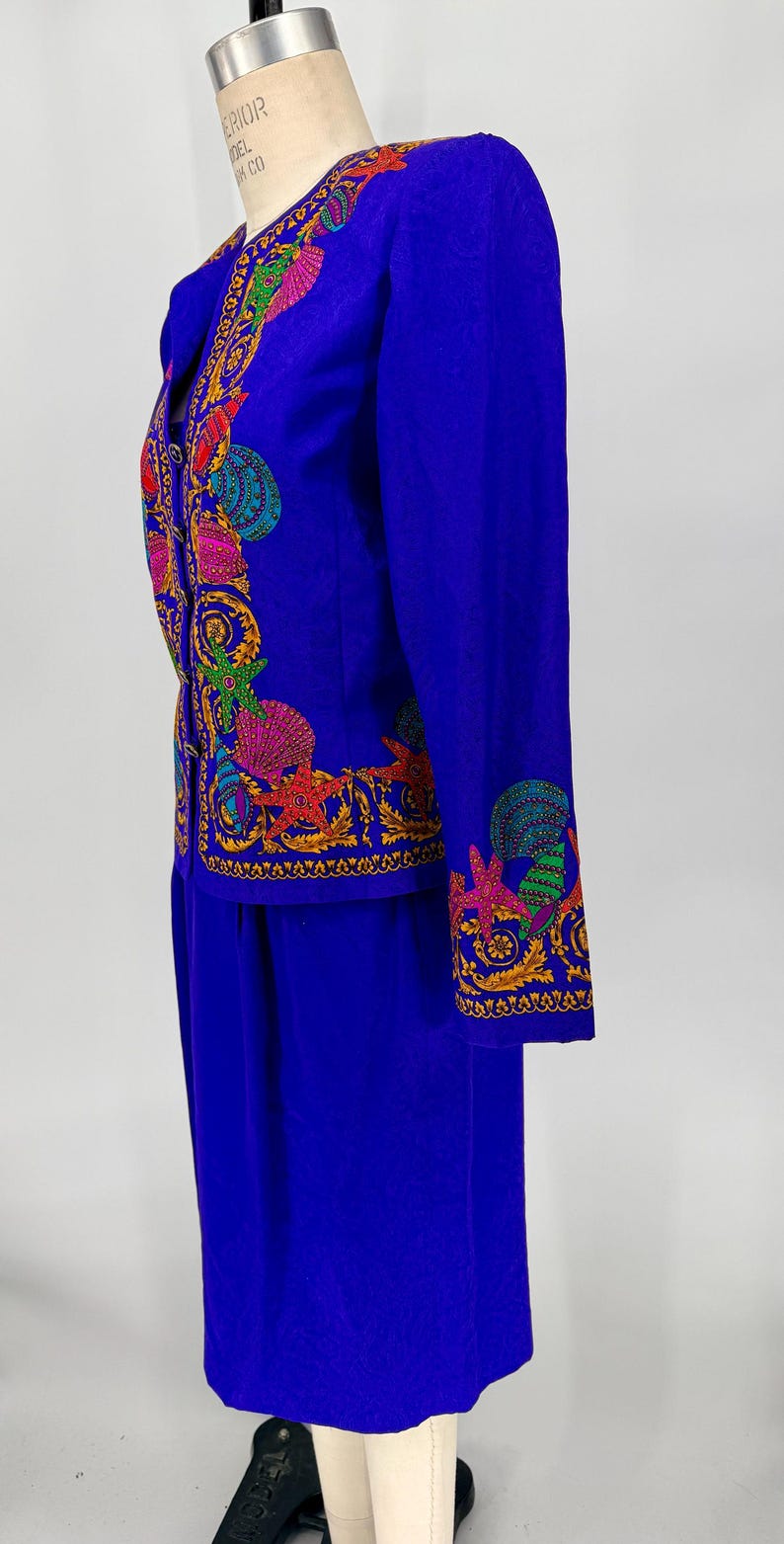 May include: A vibrant blue two-piece suit featuring intricate, colorful embroidered seashell and starfish designs. The jacket has long sleeves and a tailored fit, paired with a matching skirt. The embroidery includes shades of red, green, yellow, and pink.