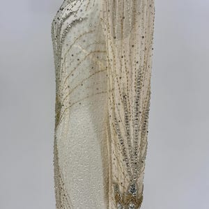 K&ouml;nnte beinhalten: Ein elfenbeinfarbenes, bodenlanges Kleid mit langen, durchsichtigen &Auml;rmeln. Das Kleid ist reich mit silbernen und goldenen Perlen verziert, wodurch ein funkelndes, vom Art D&eacute;co inspiriertes Design entsteht. Die &Auml;rmel und der Ausschnitt sind ebenfalls mit Perlenstickereien verziert.