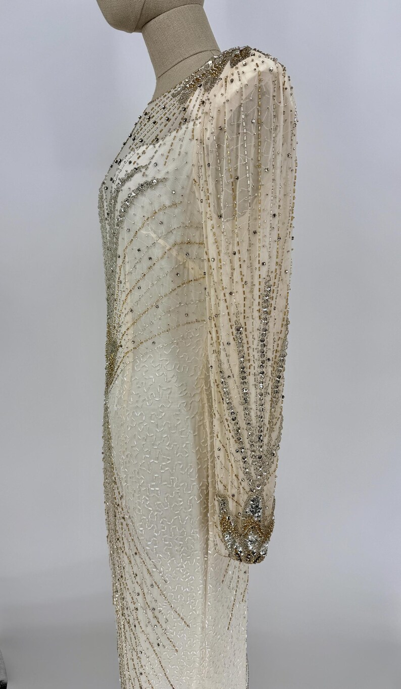 K&ouml;nnte beinhalten: Ein elfenbeinfarbenes, bodenlanges Kleid mit langen, durchsichtigen &Auml;rmeln. Das Kleid ist reich mit silbernen und goldenen Perlen verziert, wodurch ein funkelndes, vom Art D&eacute;co inspiriertes Design entsteht. Die &Auml;rmel und der Ausschnitt sind ebenfalls mit Perlenstickereien verziert.