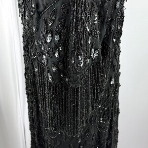 May include: A black beaded dress with a fringed hem. The dress is covered in intricate beadwork and sequins, with a detailed pattern. The sleeves are short and also feature beadwork. The dress is displayed on a mannequin.