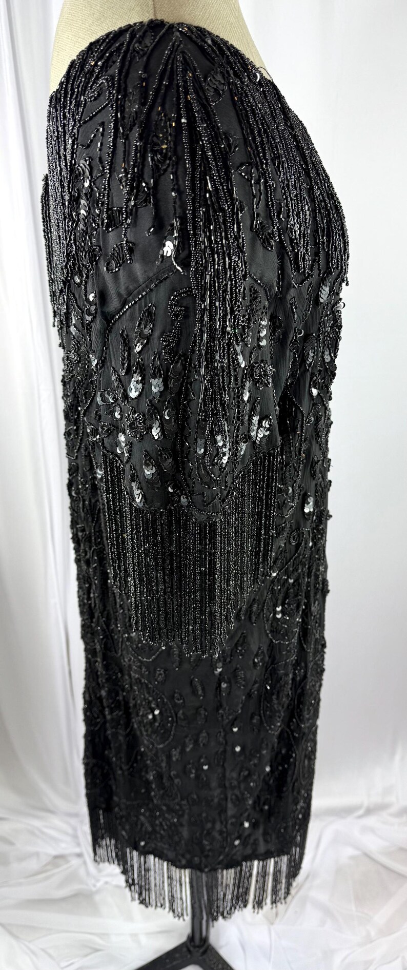 May include: A black beaded dress with a fringed hem. The dress is covered in intricate beadwork and sequins, with a detailed pattern. The sleeves are short and also feature beadwork. The dress is displayed on a mannequin.