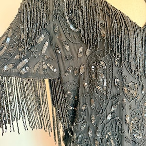 May include: A dark grey beaded top with fringe detail. The top features intricate sequin and beadwork in a paisley design. The sleeves are adorned with long beaded fringe, adding movement and texture to the garment.