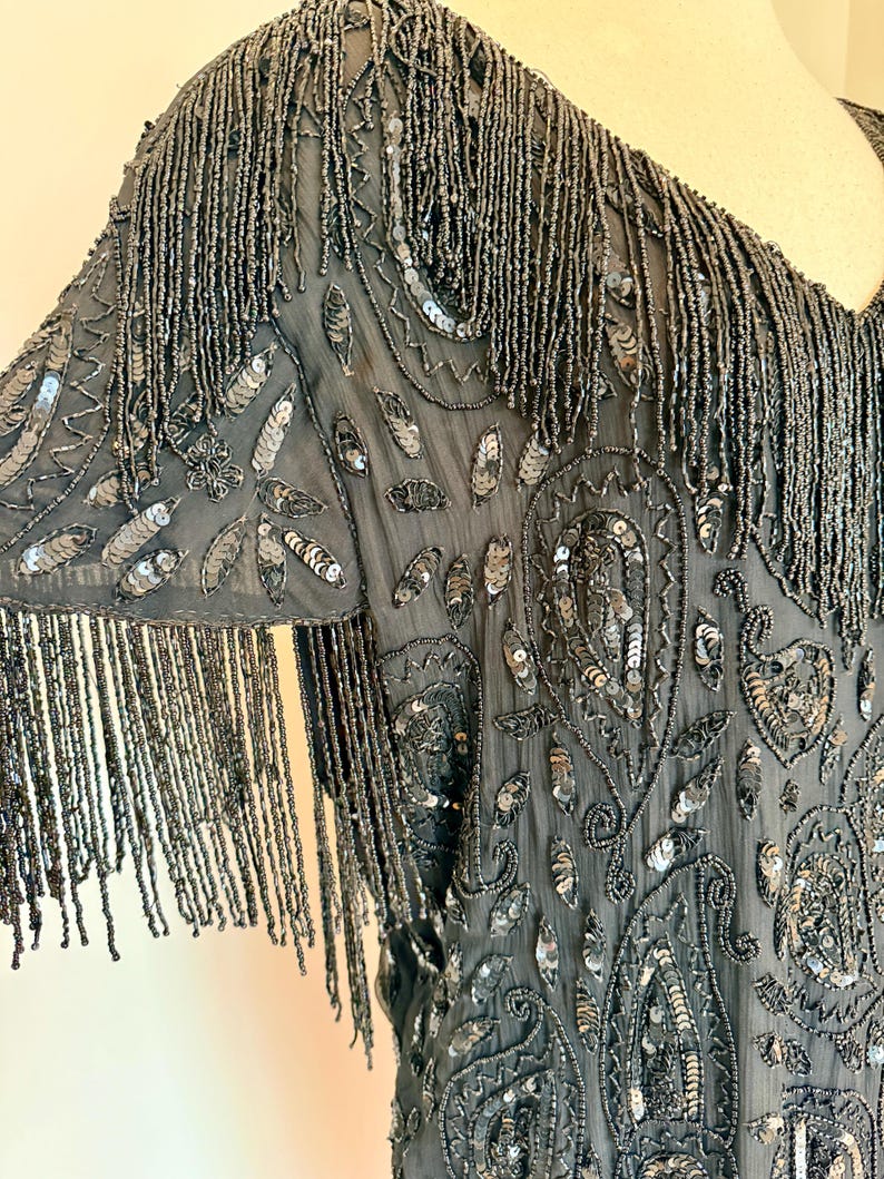 May include: A dark grey beaded top with fringe detail. The top features intricate sequin and beadwork in a paisley design. The sleeves are adorned with long beaded fringe, adding movement and texture to the garment.