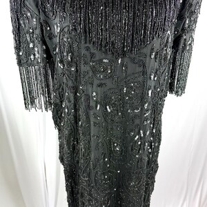 May include: Black beaded dress with a V-neck and short sleeves. The dress has a paisley pattern and fringe detailing on the neckline, sleeves, and hem. The dress is covered in sequins and beads.