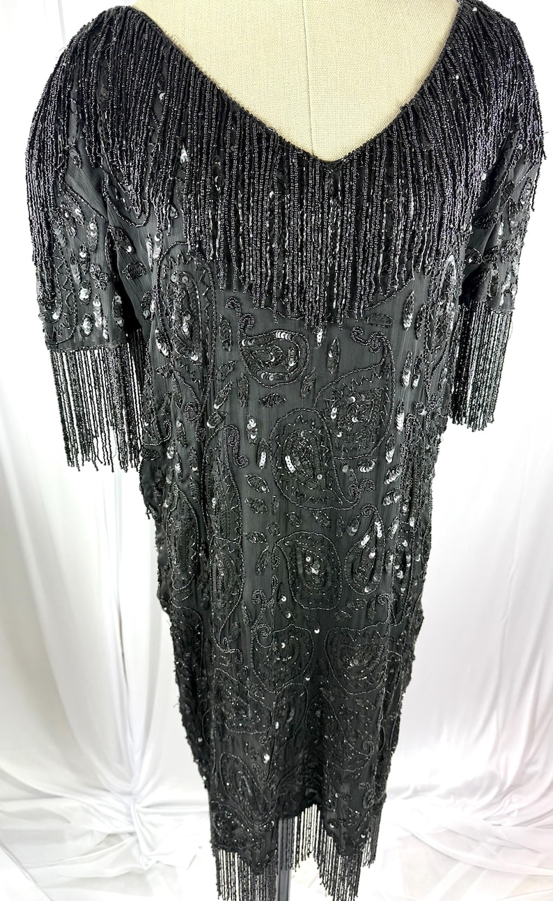 May include: Black beaded dress with a V-neck and short sleeves. The dress has a paisley pattern and fringe detailing on the neckline, sleeves, and hem. The dress is covered in sequins and beads.