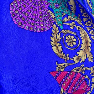 May include: Close-up of a vibrant blue fabric with a detailed pattern. The design features ornate gold flourishes, seashells in shades of pink and purple, and a green starfish, all adorned with jewel-like accents. The fabric has a subtle paisley texture.