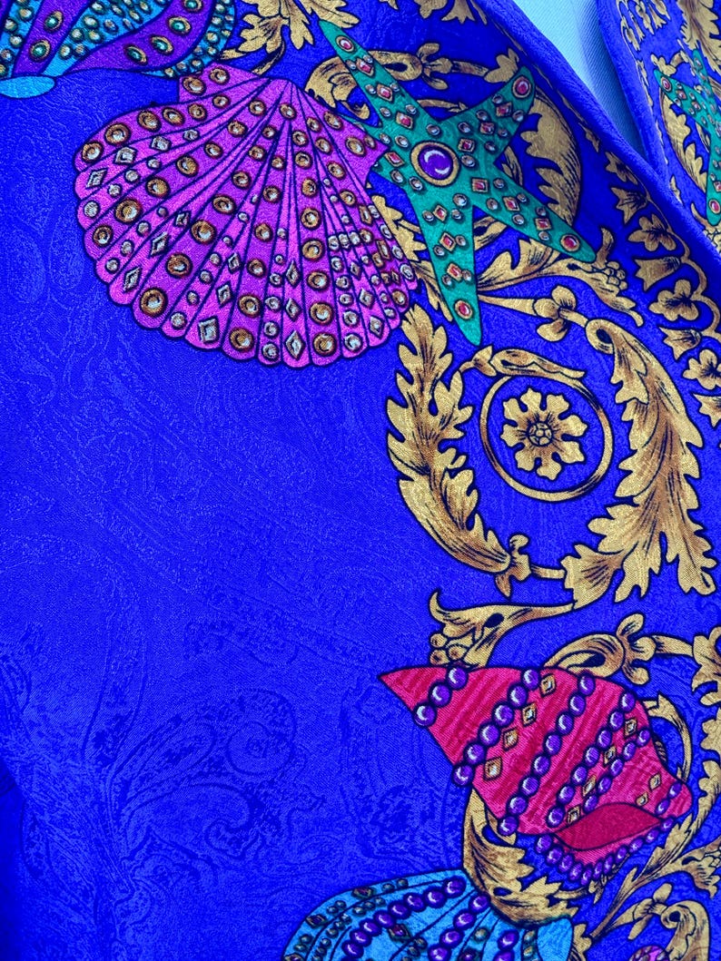 May include: Close-up of a vibrant blue fabric with a detailed pattern. The design features ornate gold flourishes, seashells in shades of pink and purple, and a green starfish, all adorned with jewel-like accents. The fabric has a subtle paisley texture.