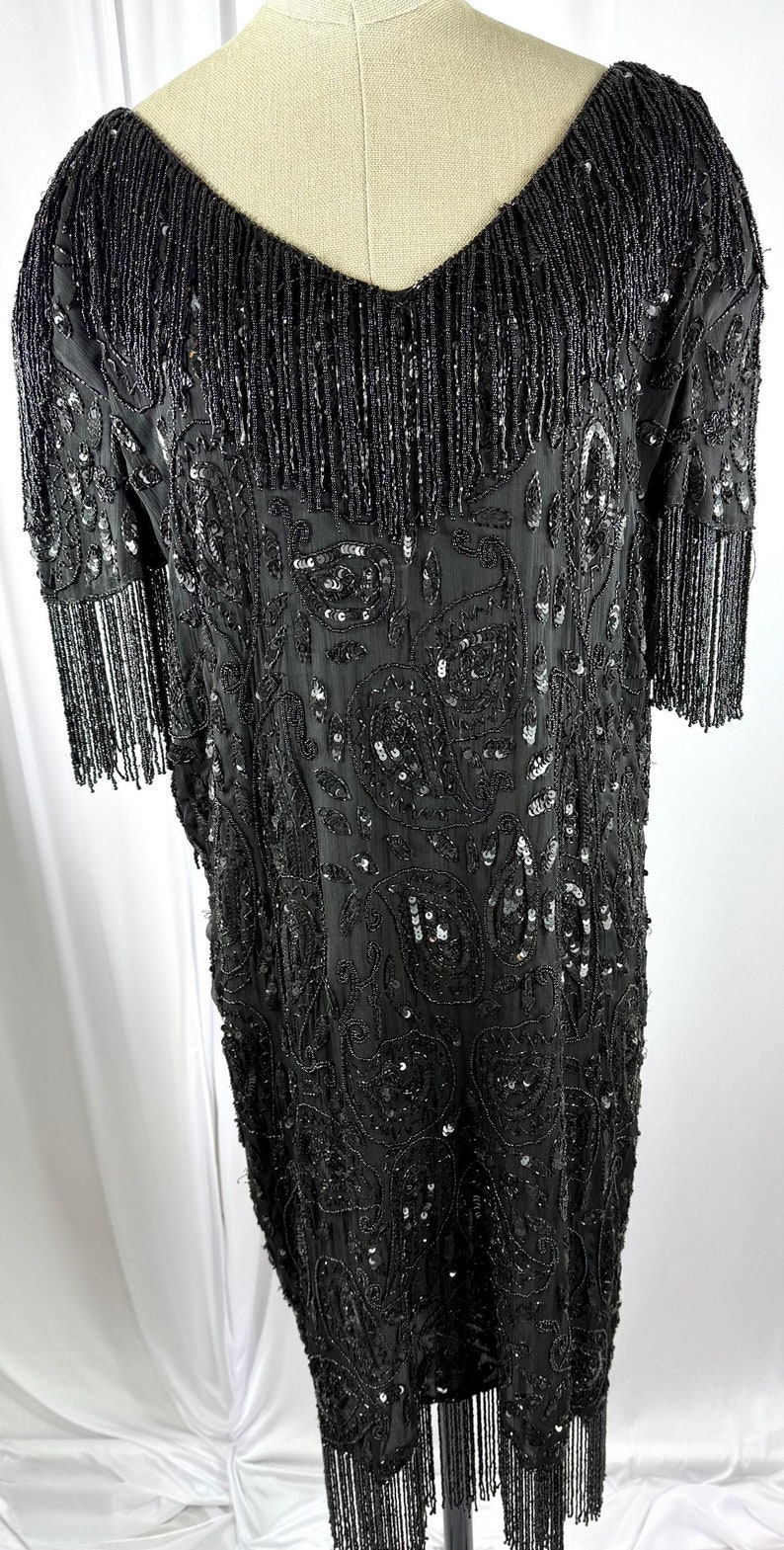 May include: A black beaded dress with a V-neck and short sleeves. The dress has a paisley pattern with sequins and fringe detailing on the sleeves and hem. The dress is displayed on a mannequin.
