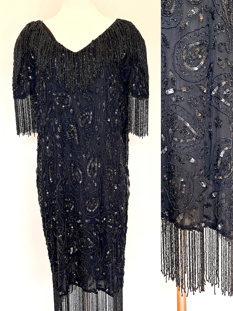 May include: A black beaded dress with a V-neck and short sleeves. The dress has a paisley pattern and fringe detailing on the neckline, sleeves, and hem. It is embellished with sequins and beads, creating a shimmering effect.