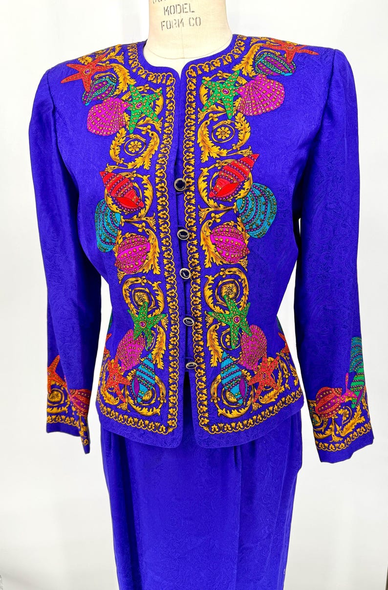May include: A vibrant blue two-piece suit featuring a jacket and skirt. The jacket is adorned with colorful embroidered seashells, starfish, and ornate gold detailing. The suit has a classic silhouette with a button-down front.
