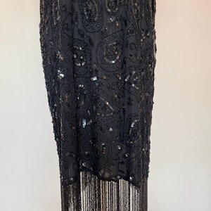 May include: A black, beaded dress with a paisley pattern and fringe detailing. The dress features intricate beadwork and sequins, with fringe along the hemline and sleeves. The dress is displayed on a wooden stand.