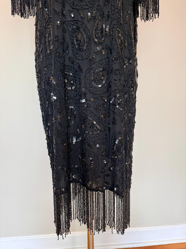 May include: A black, beaded dress with a paisley pattern and fringe detailing. The dress features intricate beadwork and sequins, with fringe along the hemline and sleeves. The dress is displayed on a wooden stand.