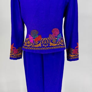 May include: A vibrant blue two-piece suit featuring intricate seashell embroidery in shades of pink, red, green, and gold. The jacket and skirt are displayed on a mannequin, showcasing the detailed design.