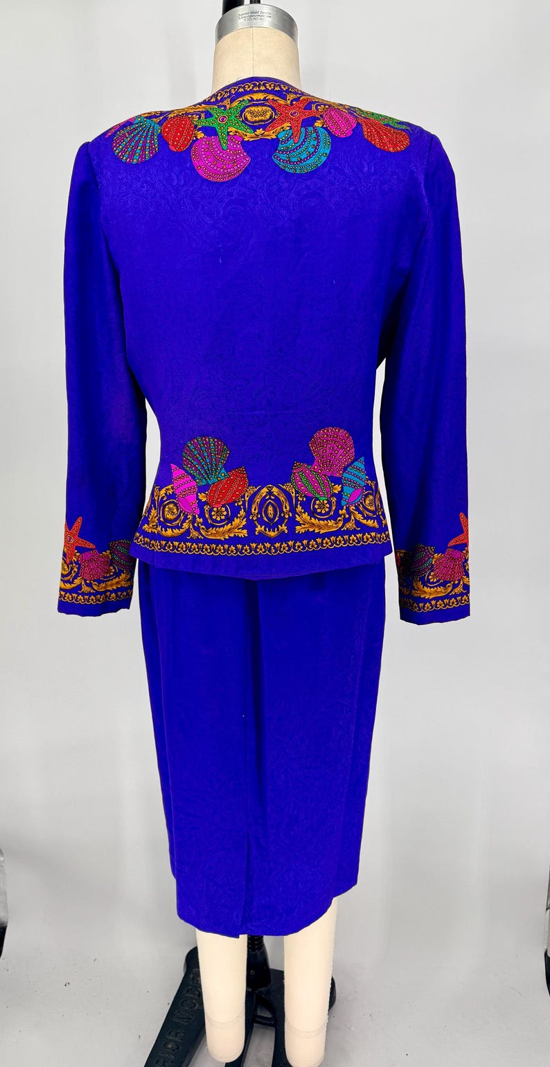 May include: A vibrant blue two-piece suit featuring intricate seashell embroidery in shades of pink, red, green, and gold. The jacket and skirt are displayed on a mannequin, showcasing the detailed design.