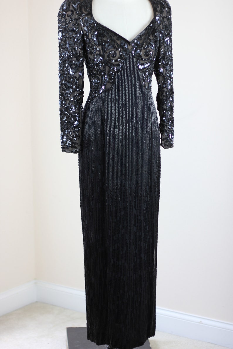 May include: A long black sequined evening gown with a V-neckline and long sleeves. The gown is fully sequined with a pattern of swirls and lines.