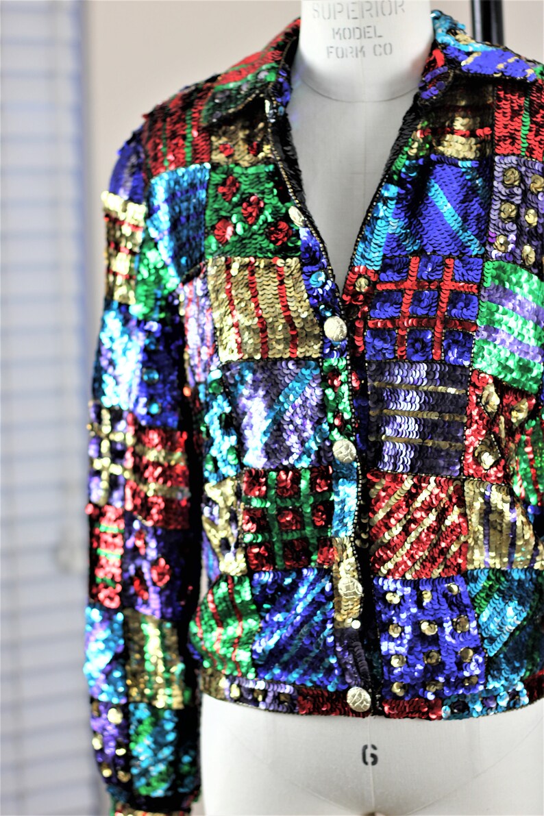 Puede incluir: Chaqueta de lentejuelas colorida con dise&ntilde;o de patchwork. La chaqueta presenta una variedad de colores, incluyendo rojo, verde, azul y dorado. Tiene cierre de cremallera y botones dorados. La chaqueta se muestra en un maniqu&iacute;.