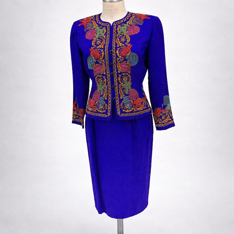 May include: A vibrant blue skirt suit with an embroidered jacket. The jacket features colorful floral and seashell designs in red, green, yellow, and purple. The skirt is a matching solid blue. The suit is displayed on a mannequin.