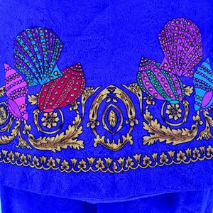 May include: Close-up of a vibrant blue fabric with a detailed pattern. The design features colorful seashell-like shapes in pink, teal, and red, accented with gold floral and scrollwork. The fabric has a paisley texture.