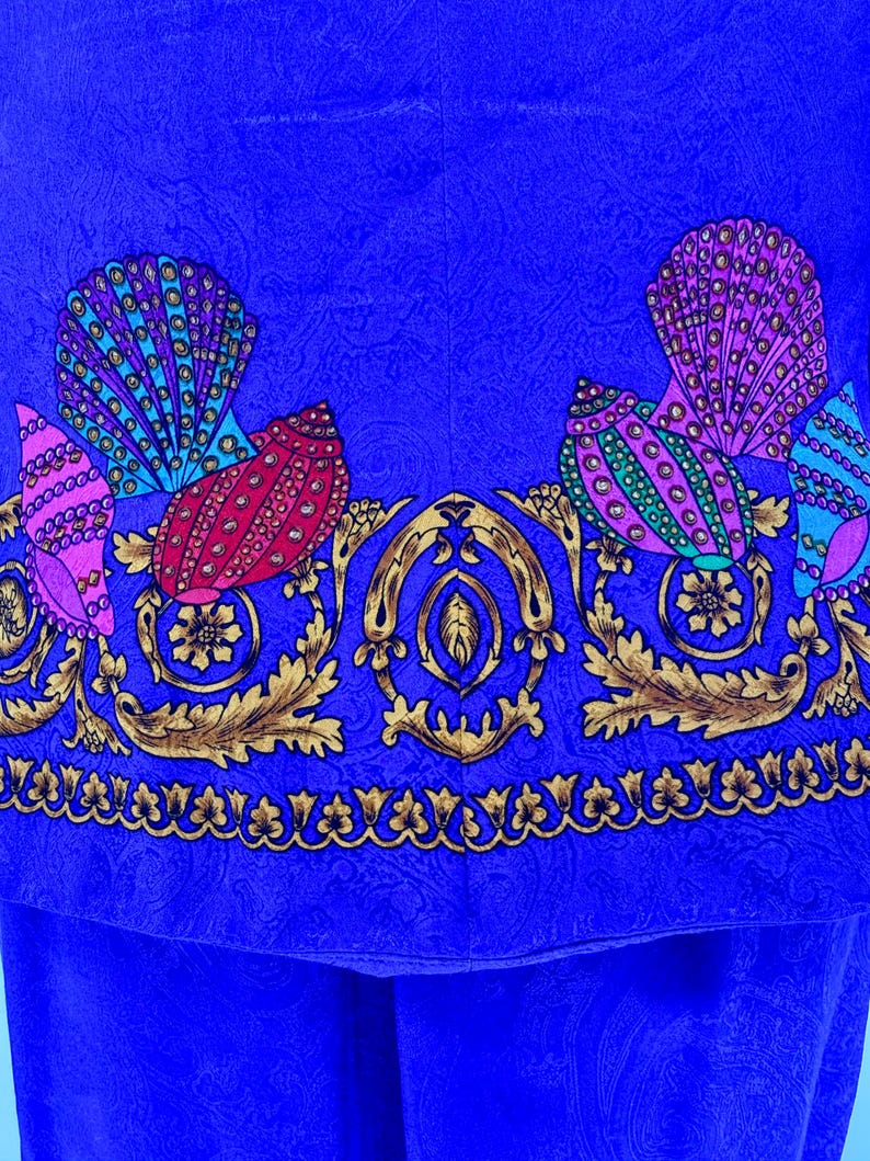 May include: Close-up of a vibrant blue fabric with a detailed pattern. The design features colorful seashell-like shapes in pink, teal, and red, accented with gold floral and scrollwork. The fabric has a paisley texture.