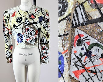 May include: A sequined cropped jacket with a round neck and long sleeves. The jacket has an abstract pattern in black, red, blue, and white on a cream base. It is displayed on a mannequin, showcasing its design and style.