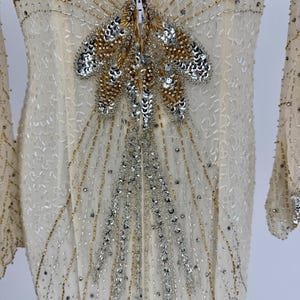 K&ouml;nnte beinhalten: Elfenbeinfarbenes Abendkleid mit langen &Auml;rmeln, verziert mit silbernen und goldenen Perlen und Pailletten. Das Oberteil hat ein zentrales Design aus Perlen und Pailletten mit Rei&szlig;verschluss. Das Kleid hat einen V-Ausschnitt und eine flie&szlig;ende Silhouette.