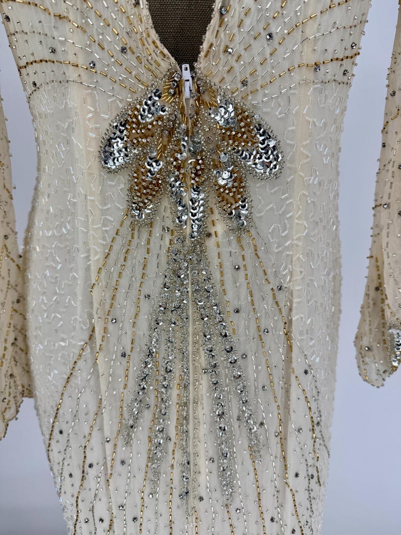 K&ouml;nnte beinhalten: Elfenbeinfarbenes Abendkleid mit langen &Auml;rmeln, verziert mit silbernen und goldenen Perlen und Pailletten. Das Oberteil hat ein zentrales Design aus Perlen und Pailletten mit Rei&szlig;verschluss. Das Kleid hat einen V-Ausschnitt und eine flie&szlig;ende Silhouette.