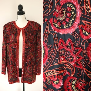 May include: A black sequined jacket with a paisley pattern in red and gold. The jacket has a long sleeve and a front closure.