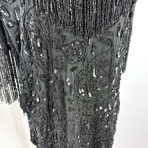 May include: Black beaded dress with a V-neck and short sleeves. The dress has detailed paisley patterns and fringe. The fabric is embellished with sequins and beads, creating a shimmering effect.