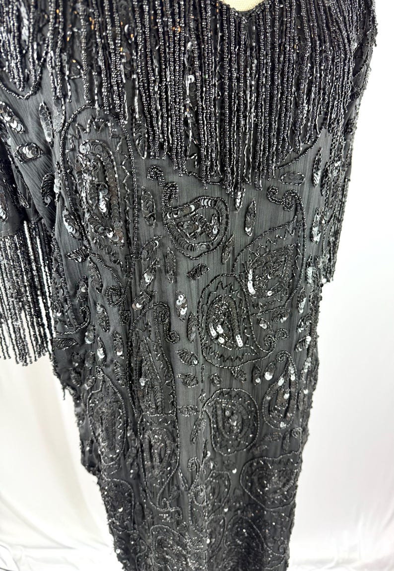 May include: Black beaded dress with a V-neck and short sleeves. The dress has detailed paisley patterns and fringe. The fabric is embellished with sequins and beads, creating a shimmering effect.