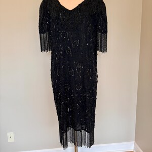 May include: A black beaded dress with a V-neck and short sleeves. The dress has fringe detailing on the sleeves and hem. The dress is displayed on a mannequin.