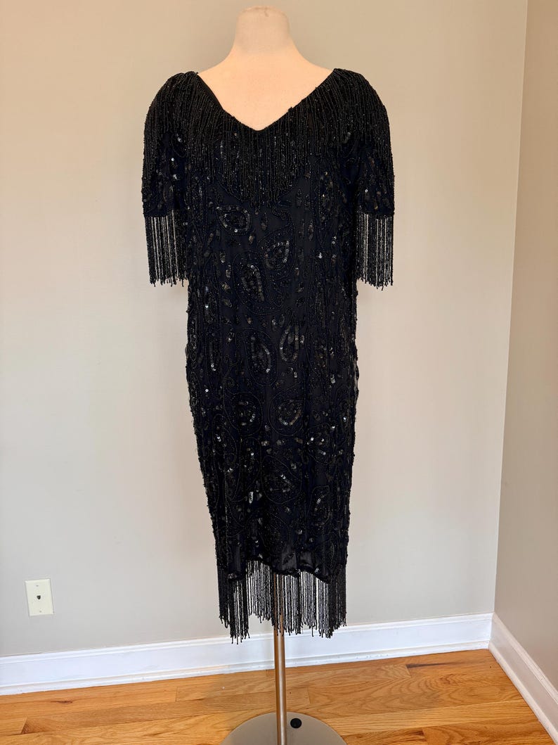 May include: A black beaded dress with a V-neck and short sleeves. The dress has fringe detailing on the sleeves and hem. The dress is displayed on a mannequin.