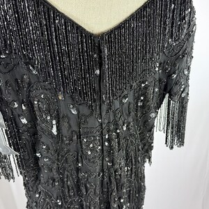 May include: A black beaded dress with a V-neck and short sleeves. The dress has detailed beadwork and sequin embellishments, with fringe detailing on the neckline and sleeves. The dress is displayed on a mannequin.