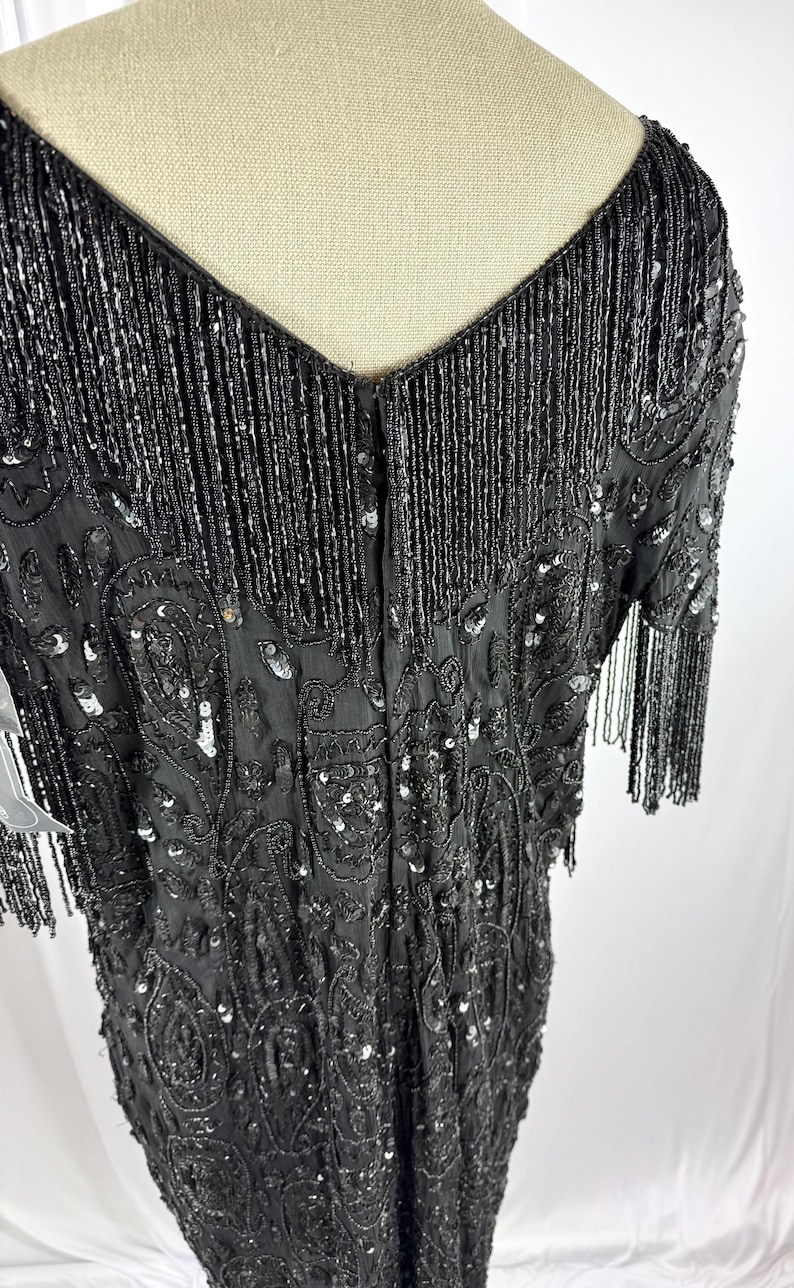 May include: A black beaded dress with a V-neck and short sleeves. The dress has detailed beadwork and sequin embellishments, with fringe detailing on the neckline and sleeves. The dress is displayed on a mannequin.